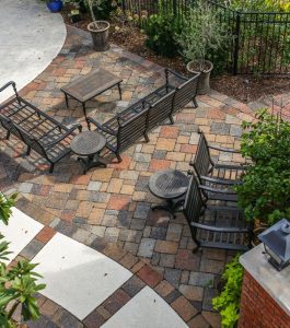 Brick patio area with metal chairs and tables next to a curved swimming pool, surrounded by plants and a fence.