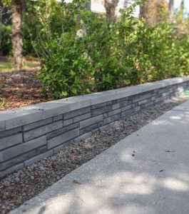 Low gray stone retaining wall borders a landscaped garden with green shrubs next to a concrete walkway.