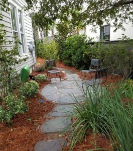 Stone walkway lined with mulch and plants, leading to two metal chairs beside a house.