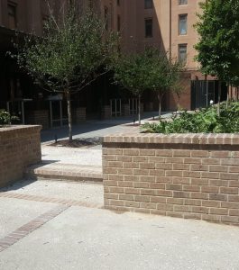 A brick courtyard features raised planters with trees, surrounded by concrete paths and brick buildings.