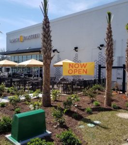 Outdoor seating area in front of a café with umbrellas and a large "Now Open" sign displayed by the entrance.