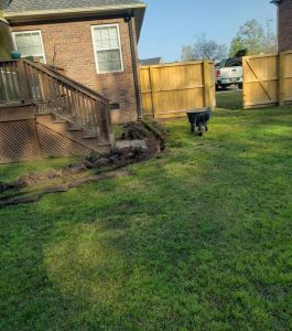 A trench is dug along a backyard deck with a wheelbarrow nearby on a grassy lawn next to a wooden fence.