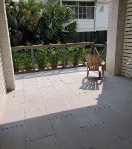 A single brown chair is placed on a tiled patio, shaded and facing a railing with greenery and a building in the background.