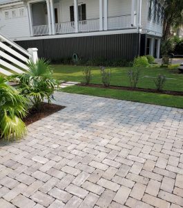 A paved stone walkway leads to a lawn with trimmed shrubs in front of a large white house with a porch.