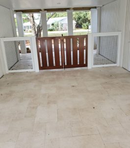 Open-air covered patio with tiled flooring, white walls, and a wooden gate with wire fencing, facing a yard.