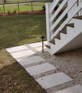 Stone pavers lead to a staircase with a white railing beside a grassy lawn and a small garden light.