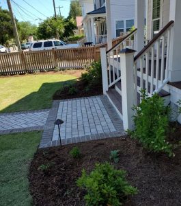 Front yard of a house with a brick walkway, steps, garden beds, small shrubs, and a wooden fence.