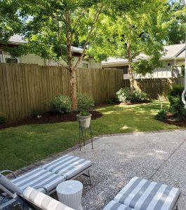Backyard with a patio, two striped lounge chairs, green lawn, trees, shrubs, and a wooden privacy fence.