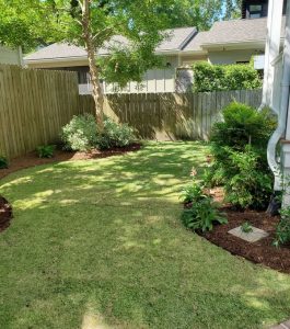 A small backyard with green grass, landscaped flower beds, a wooden fence, and trees providing shade.