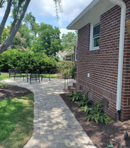 Brick house with a paved walkway leading to a patio area with black metal chairs and a table in a landscaped yard.