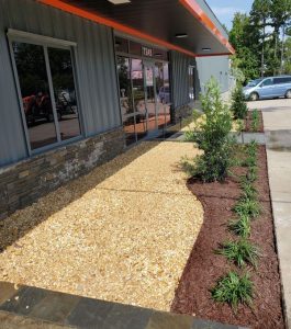 Gravel walkway and mulched garden beds with shrubs run along the exterior of a commercial building with large windows.