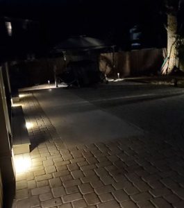 Brick patio at night with ground lighting along the edge and a fenced yard in the background.