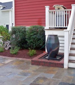 A large ceramic water feature stands by stairs next to shrubs and mulch in a neatly landscaped yard with stone paving.