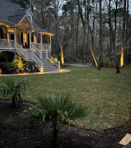 A house with a large porch and stairs is shown at dusk, with exterior lighting illuminating the yard and trees.