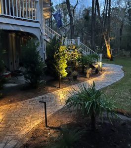 Curved stone pathway with outdoor lights leads to a house with stairs, surrounded by trees and landscaping at dusk.