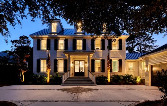 Two-story house with dormer windows, dark shutters, and a lit entrance, photographed at dusk with a driveway in front.