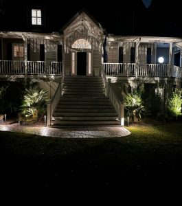Two-story house with wide front steps is illuminated at night by outdoor lights highlighting the porch and plants.
