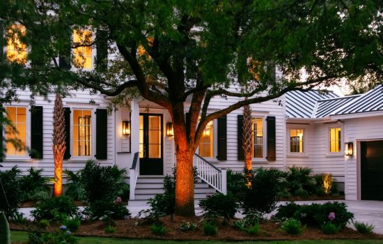 White house with black shutters and a front porch, illuminated at dusk, with trees and landscaping in the foreground.