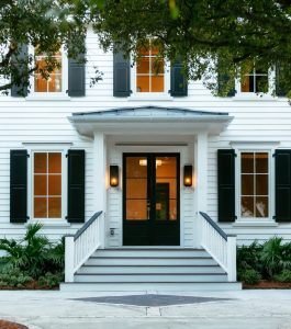 Two story home with white siding and black trim featuring plants and trees in front