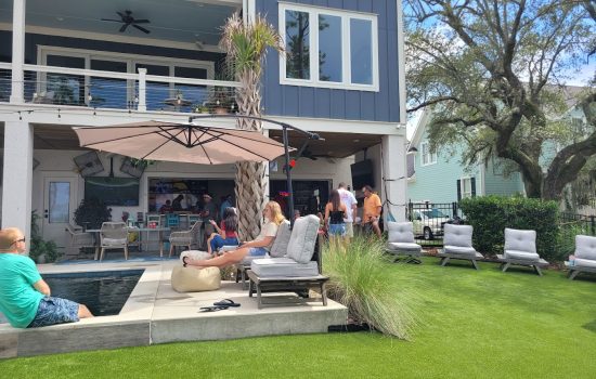 People socializing by a pool in a backyard with outdoor seating, a large umbrella, and artificial grass on a sunny day.