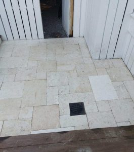 Outdoor patio with light stone tiles, a central drain, wooden steps, and white paneled walls.