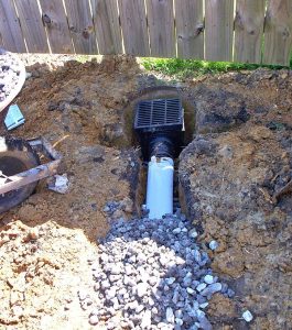 A drainage system is being installed in a yard with a grate, pipe, and surrounding gravel visible amid dug-up soil.