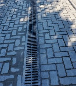 A paved surface with interlocking bricks and a central metal drainage grate running vertically.
