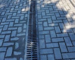 A paved surface with interlocking bricks and a central metal drainage grate running vertically.
