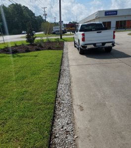A white pickup truck is parked next to a driveway bordered by grass and a strip of gravel on a sunny day.