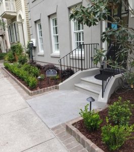 A concrete walkway leads to a gray building entrance with stairs, black railing, and landscaped garden beds.