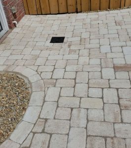 A paved patio with light-colored stone tiles, a small drainage grate, gravel area, and a wooden fence in the background.