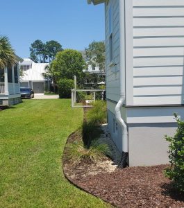 Side view of a white house with landscaped yard, green grass, mulch, and a visible downspout on a sunny day.