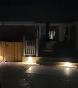 A backyard patio at night with ground lights illuminating a fence, walkway, and seating area near a house.