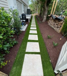 A straight pathway of square stone pavers set in artificial grass runs alongside a house with plants and mulch on both sides.