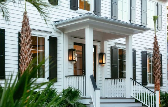 Front entrance of a white house with black shutters, porch columns, steps, and palm trees in the landscaping.