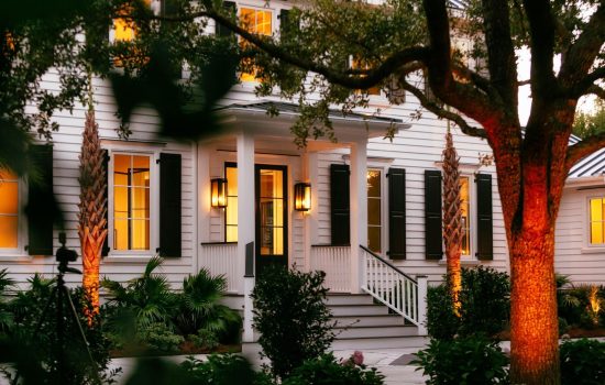 White two-story house with black shutters, lit windows, a front porch, and landscaping at dusk.