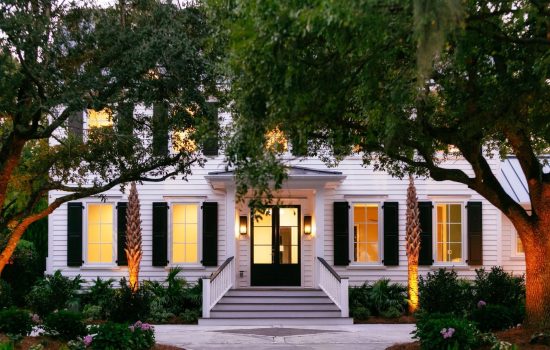 White two-story house with black shutters, front steps, and warm lights, surrounded by trees and landscaping.