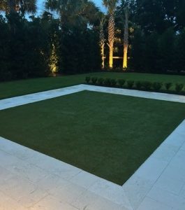 A square patch of grass is bordered by light stone tiles, with trees and shrubs in the background illuminated by garden lights.