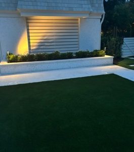 A modern house exterior with a lit planter, green lawn, white paving stones, and horizontal window shutters at dusk.