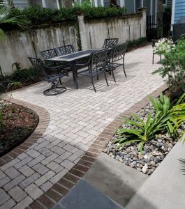 Outdoor patio with metal table and chairs on light brick pavers, bordered by plants, rocks, and a stone pathway.