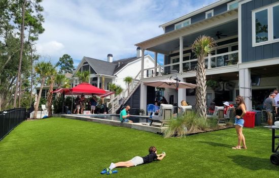 People gather in a backyard with artificial turf, pool, and umbrellas near a modern house on a sunny day.