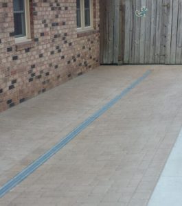 A brick driveway with a rectangular drainage grate runs alongside a brick house, ending at a wooden gate.