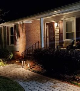 Single-story brick house at night with a lit front porch, two chairs, and a stone walkway bordered by shrubs.