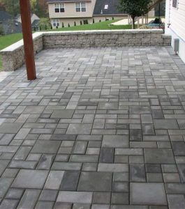 Backyard patio with gray interlocking pavers, bordered by a low stone wall and adjacent to a house siding.