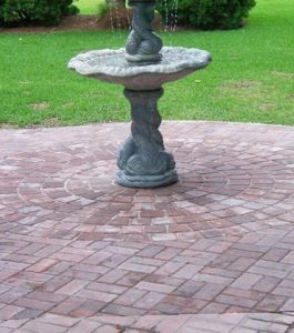 A decorative stone birdbath stands on a circular brick patio surrounded by green grass.