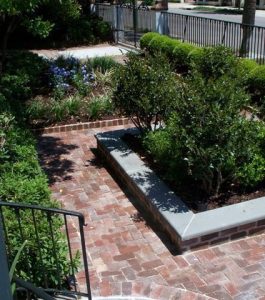 Brick walkway and steps lead to a raised garden bed with shrubs and surrounding greenery near a street.