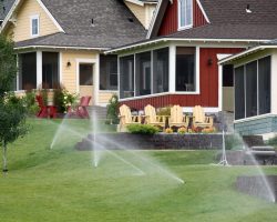 Lawn sprinklers watering grass in front of colorful houses with porches and outdoor chairs.