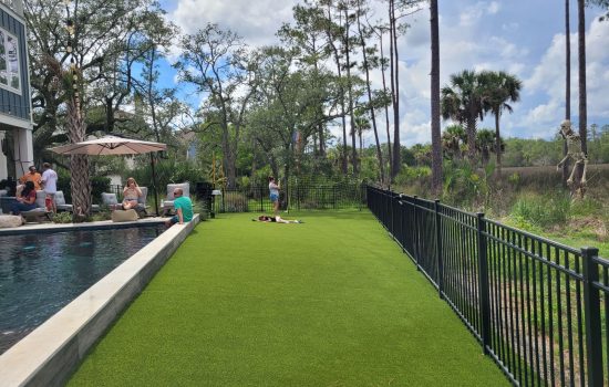 People relax by a pool and on artificial grass near a black fence, with trees and greenery in the background.