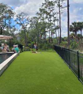 People relax by a pool and on artificial grass near a black fence, with trees and greenery in the background.